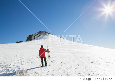 Male skier standing on alpine slope holding skis and pole, wearing red jacket, footprints trailing 135715972