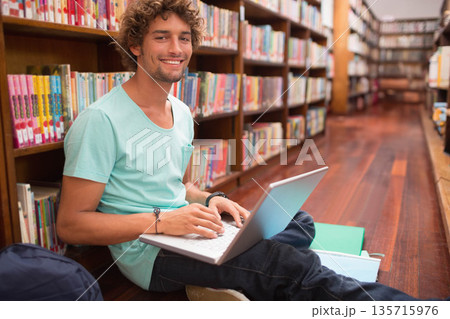 Man in twenties sitting on wooden floor between bookshelves in library typing on silver laptop Man in twenties sitting on wooden floor between bookshelves in library typing on silver laptop 135715976