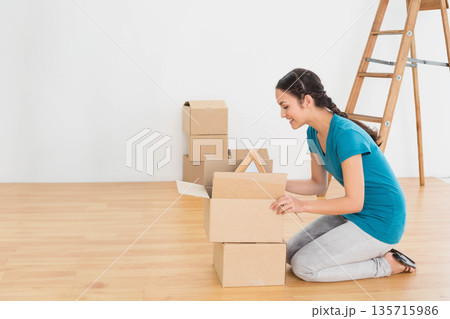 Woman kneeling on floor in white room opening boxes, handling frame near ladder, copy space Woman kneeling on floor in white room opening boxes, handling frame near ladder, copy space 135715986