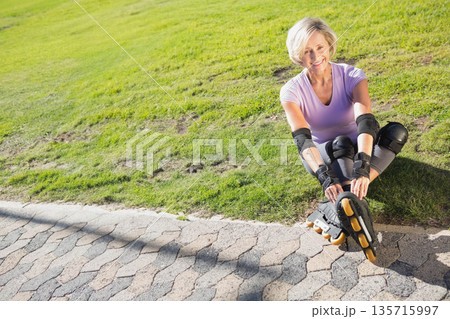 Senior woman sitting at park in lavender fixing skates with yellow wheels and pads, copy space Senior woman sitting at park in lavender fixing skates with yellow wheels and pads, copy space 135715997