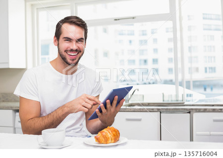 Man sitting at kitchen table wearing white T-shirt, holding blue tablet, smiling near coffee cup Man sitting at kitchen table wearing white T-shirt, holding blue tablet, smiling near coffee cup 135716004