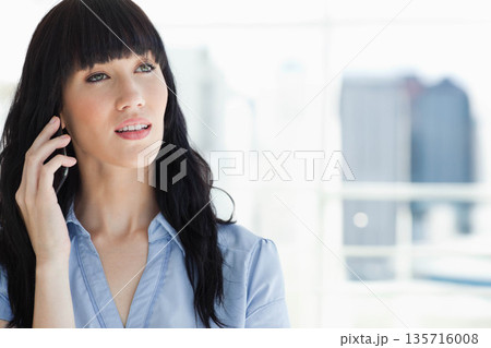 Adult woman holding phone to ear, looking upward near office window in collared blouse, copy space Adult woman holding phone to ear, looking upward near office window in collared blouse, copy space 135716008