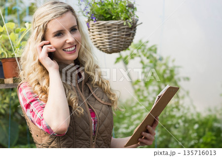 Woman in greenhouse wearing brown vest, using phone, holding clipboard among plants, hanging basket Woman in greenhouse wearing brown vest, using phone, holding clipboard among plants, hanging basket 135716013