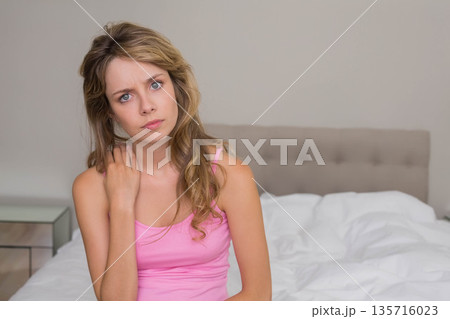 Adult female sitting on bed, touching neck and looking toward camera in pink tank top 135716023