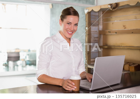 Mid 30s female standing at cafe counter wearing white shirt holding takeaway cup and using laptop Mid 30s female standing at cafe counter wearing white shirt holding takeaway cup and using laptop 135716025