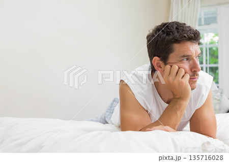 Mid-adult man lying on bed in white tank and patterned pajama pants near window, copy space 135716028