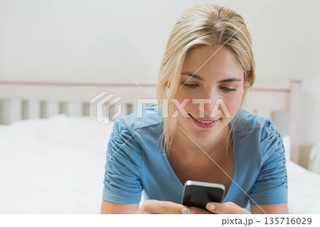 Female in twenties reclining on white bedding in bedroom, holding smartphone and wearing blue top 135716029