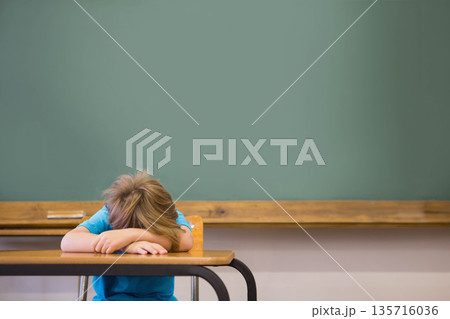 Boy child resting head on arms at desk in classroom with green chalkboard, wooden chalk tray Boy child resting head on arms at desk in classroom with green chalkboard, wooden chalk tray 135716036