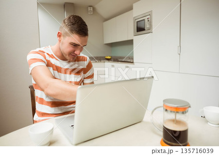 Male sitting at kitchen table working on silver laptop in orange striped t-shirt with French press Male sitting at kitchen table working on silver laptop in orange striped t-shirt with French press 135716039