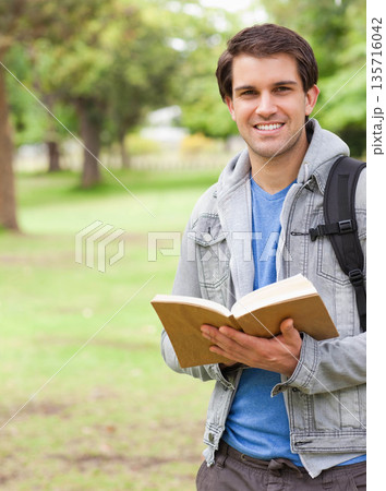 Man holding open hardcover book at chest in park, wearing denim jacket, backpack strap, copy space 135716042