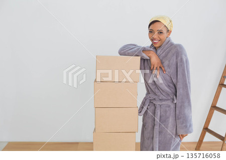 African American woman wearing robe, headscarf, leaning on boxes in room near ladder, copy space African American woman wearing robe, headscarf, leaning on boxes in room near ladder, copy space 135716058