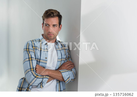 Man leaning against wall in studio, arms crossed, wearing plaid shirt and white T-shirt, copy space Man leaning against wall in studio, arms crossed, wearing plaid shirt and white T-shirt, copy space 135716062