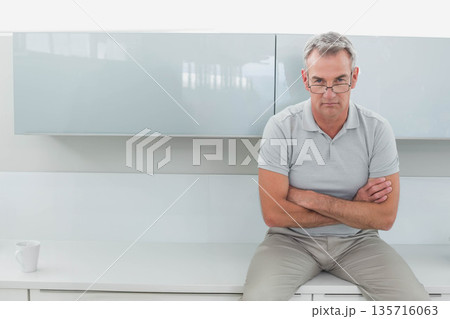 Man sitting on white counter in kitchen with white mug, crossing arms, wearing glasses, copy space Man sitting on white counter in kitchen with white mug, crossing arms, wearing glasses, copy space 135716063