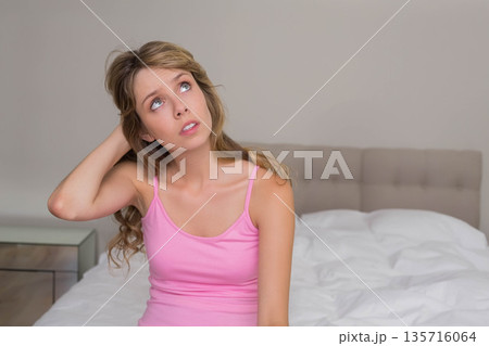 Woman in her 20s sitting on bed touching hair and looking up in bedroom with nightstand Woman in her 20s sitting on bed touching hair and looking up in bedroom with nightstand 135716064
