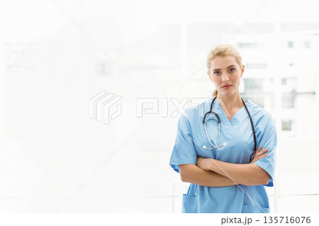 Female nurse standing with arms crossed at clinic by windows in scrubs and stethoscope, copy space 135716076