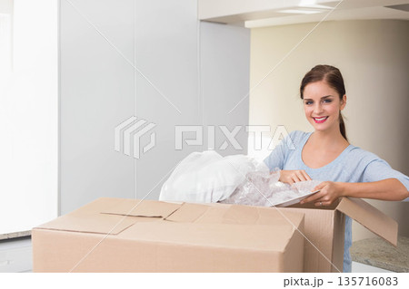 Adult female removing pillow in bubble wrap from cardboard box on kitchen island wearing blue top 135716083