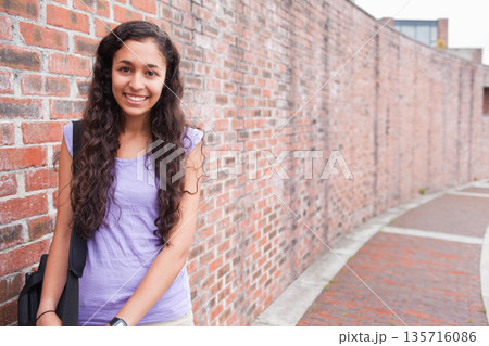 Woman in mid-20s standing, leaning on red brick wall by curved walkway, backpack strap, copy space Woman in mid-20s standing, leaning on red brick wall by curved walkway, backpack strap, copy space 135716086