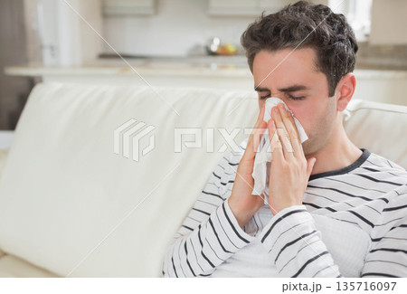 Man in twenties sitting on pale sofa holding tissue to nose, wearing striped shirt, copy space 135716097