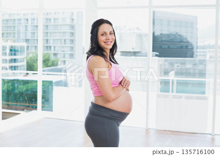 Pregnant woman standing by tall windows in bright studio cradling belly in maternity leggings Pregnant woman standing by tall windows in bright studio cradling belly in maternity leggings 135716100