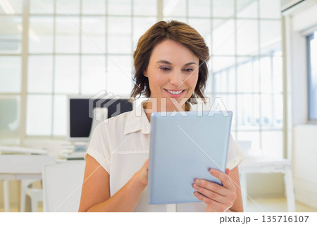 Mature woman holding tablet while standing in white blouse at open-plan office near grid windows Mature woman holding tablet while standing in white blouse at open-plan office near grid windows 135716107