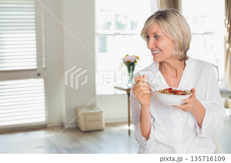 Senior woman holding cereal bowl with spoon at window in button-up smiling looking left, copy space 135716109