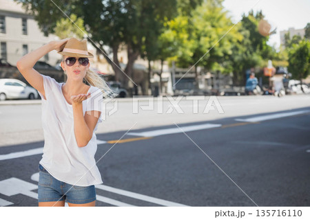 Woman 20s standing crosswalk holding hat blowing kiss in shades white tee jean shorts, copy space Woman 20s standing crosswalk holding hat blowing kiss in shades white tee jean shorts, copy space 135716110