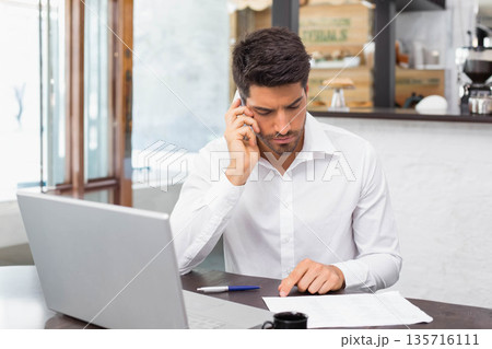 Late 20s man in white shirt sitting at cafe reading documents with laptop, phone, blue-pen, coffee 135716111