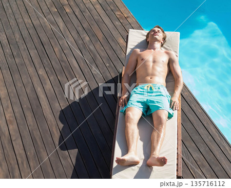Man reclining on lounge chair on wooden deck beside turquoise pool wearing teal trunks, copy space 135716112