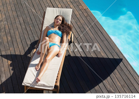 Woman in her twenties sunbathing on wooden chaise on deck by turquoise pool in blue bikini Woman in her twenties sunbathing on wooden chaise on deck by turquoise pool in blue bikini 135716114