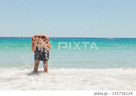 Man bending, splashing seawater onto face in shallow foamy surf at sandy beach wearing dark trunks Man bending, splashing seawater onto face in shallow foamy surf at sandy beach wearing dark trunks 135716124