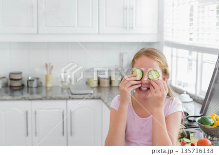 Female child holding cucumber slices over eyes laughing in bright kitchen with peppers, copy space Female child holding cucumber slices over eyes laughing in bright kitchen with peppers, copy space 135716138
