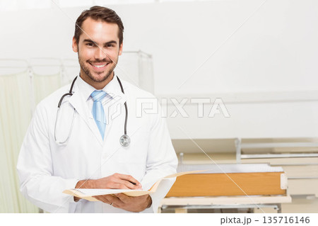 Male physician wearing lab coat, stethoscope, reading tan chart in ward with stretcher, copy space 135716146