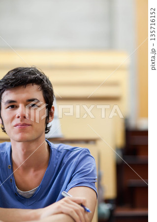 Male in twenties sitting at lecture hall desk wearing blue short-sleeve shirt and holding pen 135716152