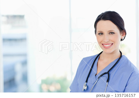 Adult woman clinician standing on right near window wearing blue scrubs and stethoscope, copy space 135716157