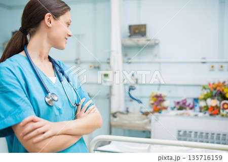 Female nurse standing arms crossed in light blue scrubs and stethoscope by bed rail, copy space 135716159