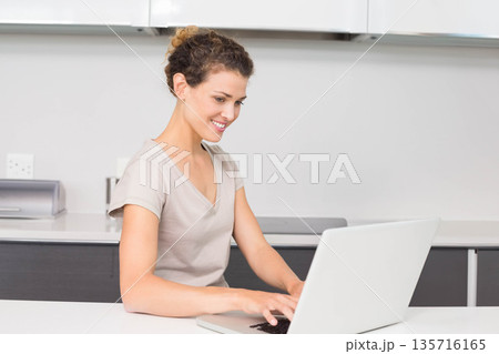 Adult woman wearing light beige T-shirt sitting at home kitchen countertop typing on silver laptop 135716165