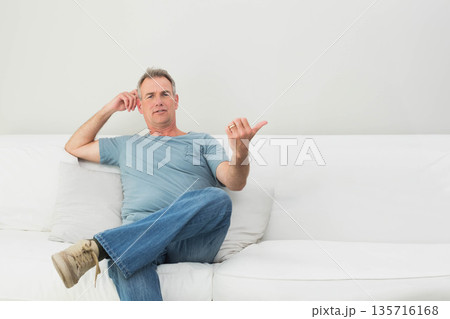 Middle aged male sitting on white sofa in minimalist living room wearing jeans, showing ring 135716168