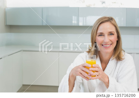 Mature woman smiling and holding glass of orange juice on countertop in bathrobe, copy space Mature woman smiling and holding glass of orange juice on countertop in bathrobe, copy space 135716200