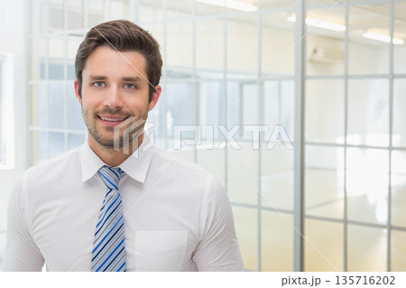 Man in his 30s standing in office with glass partitions wearing white shirt blue-striped tie 135716202