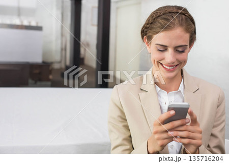 Woman sitting on bench in office wearing beige blazer and white top holding smartphone, copy space 135716204