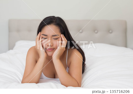Asian woman lying on stomach on bed wearing white camisole, cupping temples with pillows nearby 135716206