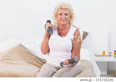 Senior woman sitting on bed in white tank top lifting small dumbbells near nightstand meds Senior woman sitting on bed in white tank top lifting small dumbbells near nightstand meds 135716226