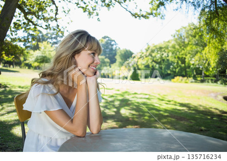 Woman sitting at round outdoor table on chair in park, wearing white short-sleeve dress, smiling Woman sitting at round outdoor table on chair in park, wearing white short-sleeve dress, smiling 135716234