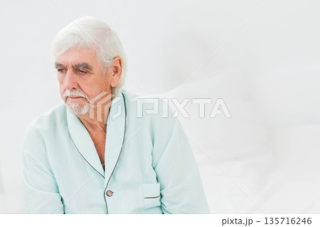 Senior man sitting on bed in bright bedroom wearing seafoam blue robe, touching white pillows 135716246