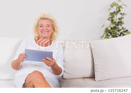 Senior woman sitting on sofa in living room holding tablet wearing white outfit, copy space 135716247