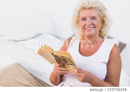 Senior female sitting on bed with beige blanket wearing top holding paperback, smiling, copy space 135716248