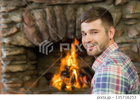 Adult bearded man smiling, sitting by stone fireplace burning logs, wearing plaid shirt, copy space 135716253