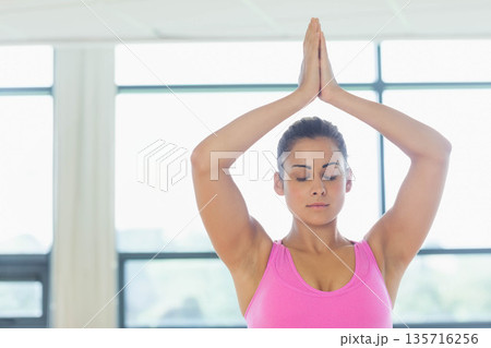 Female doing yoga with arms raised, wearing pink athletic tank top in bright studio with windows 135716256