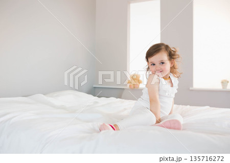Female toddler sitting on white bed in bedroom with teddy bear wearing white top, copy space Female toddler sitting on white bed in bedroom with teddy bear wearing white top, copy space 135716272