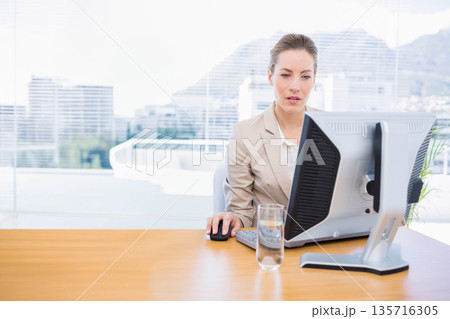 Mid adult woman in beige blazer working at office desk with monitor, mouse, glass of water Mid adult woman in beige blazer working at office desk with monitor, mouse, glass of water 135716305